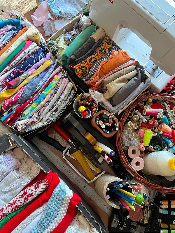 Vista cenital de un taller de costura artesanal con telas organizadas en cajas, hilos de colores en cestas de mimbre, lápices y tijeras de corte.