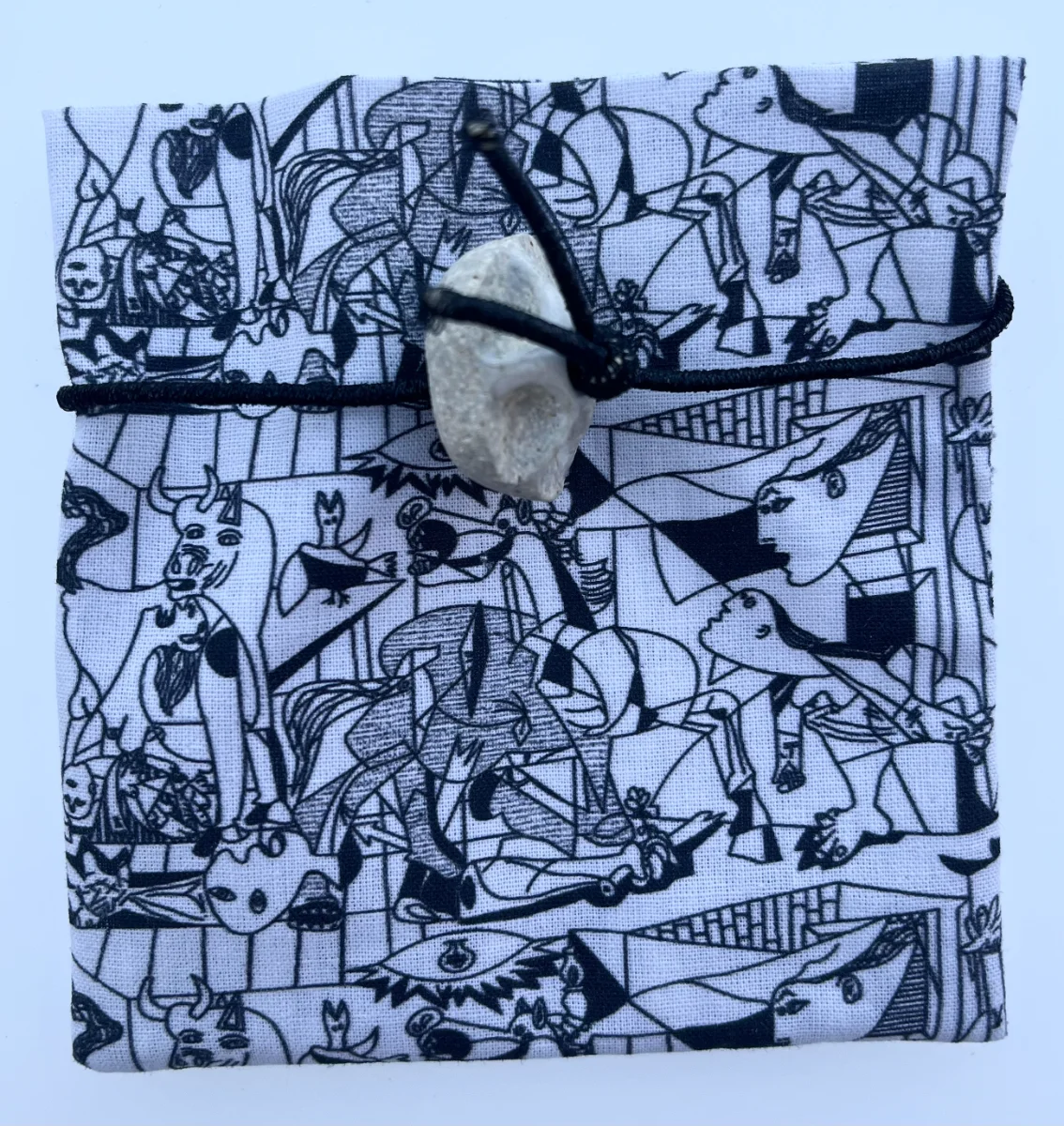 Envoltorio de tela de algodón con estampado en blanco y negro del Guernica de Picasso, atado en el centro con goma negra y pieza de cerámica esmaltada.