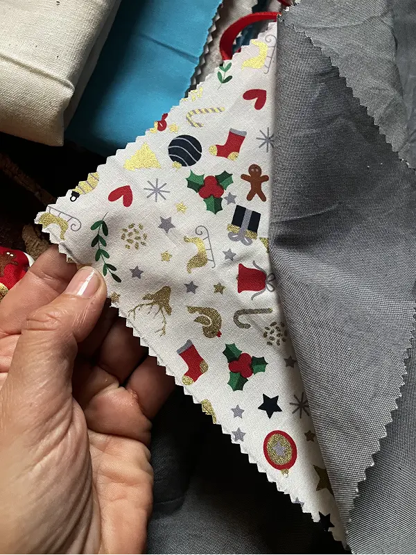 Mano sosteniendo retales de tela con borde en zigzag, destacando un estampado festivo navideño con galletas de jengibre, muérdago y estrellas.