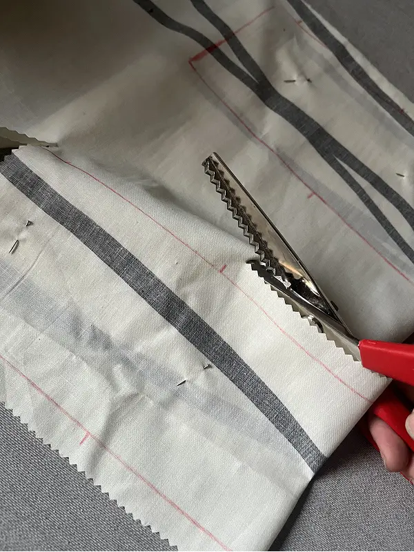 Tijeras de confección dentadas realizando un corte en zigzag sobre una tela de algodón a rayas grises y blancas.