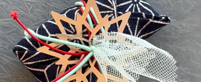 Pequeño regalo envuelto con tela de color azul marino con patrón de asanoha blanco, decorado con un copo de nieve de madera cortado con láser y cintas de color turquesa y rojo sobre una superficie gris.