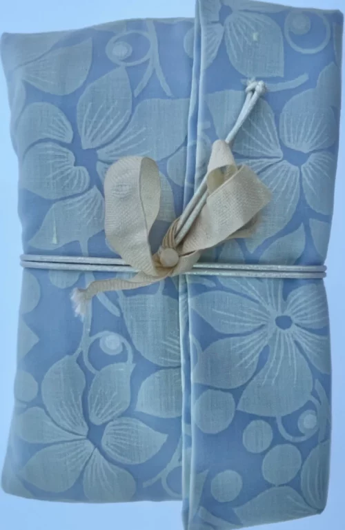 Paquete plano y rectangular envuelto en tela azul con flores blancas y cruzado con un cordón blanco y lazada beige.