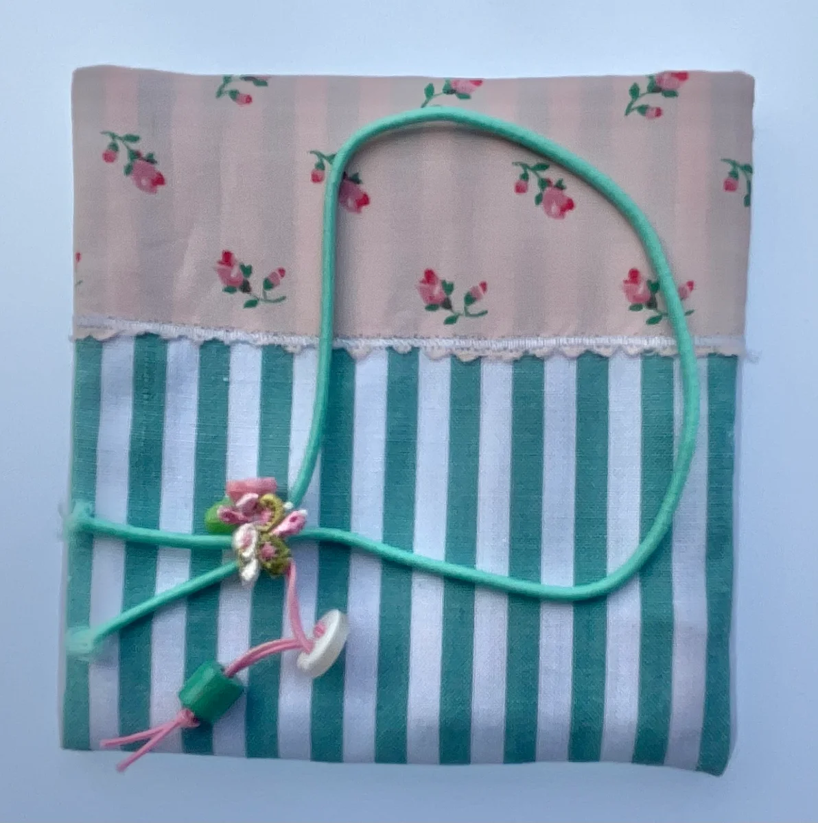 Sobre de regalo de tela combinada: parte superior rosa con pequeñas flores y parte inferior a rayas verdes y blancas, atado con cordón verde turquesa.