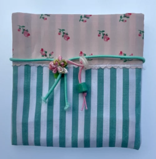Sobre de regalo de tela combinada: parte superior rosa con pequeñas flores y parte inferior a rayas verdes y blancas, atado con cordón verde turquesa.