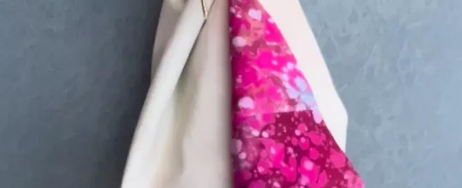 Regalo vertical envuelto en tela beige suave dejando ver un pliegue interior con estampado fucsia multicolor, atado con un botón decorativo.