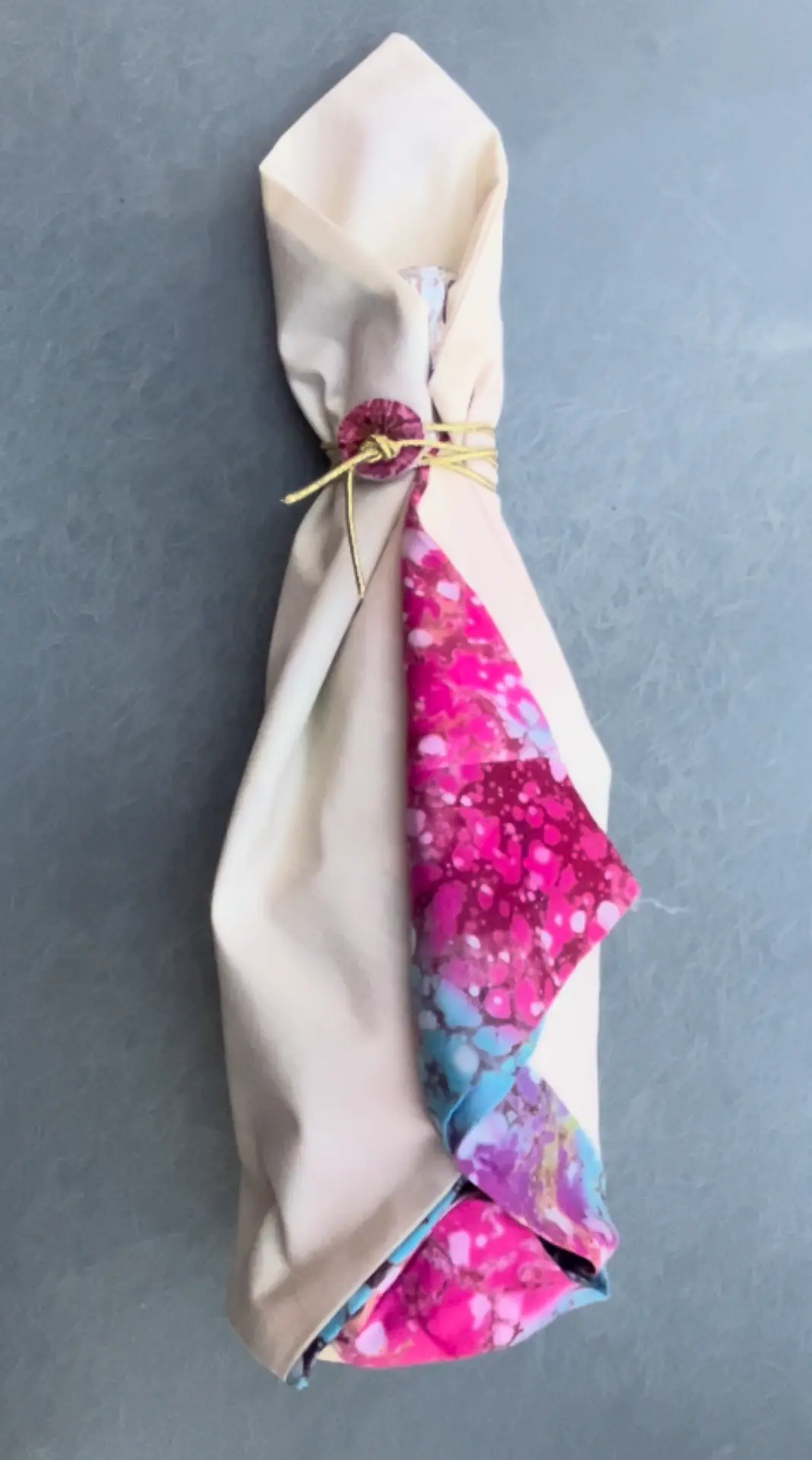 Regalo vertical envuelto en tela beige suave dejando ver un pliegue interior con estampado fucsia multicolor, atado con un botón decorativo.