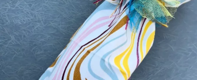 Regalo alargado envuelto en tela con estampado de ondas multicolores y atado en el cuello con una lazada de tul azul turquesa.