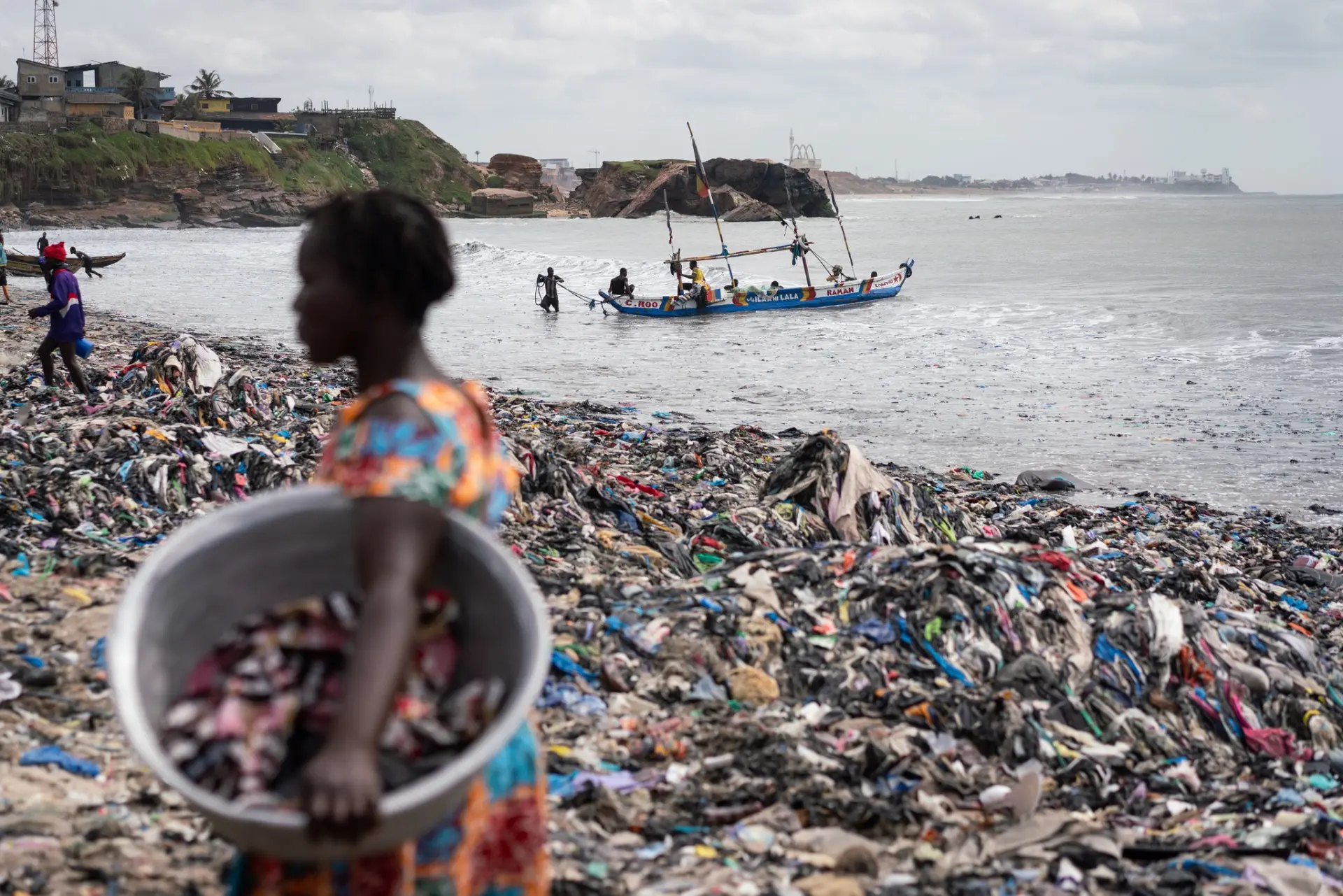 Impacto de los residuos plásticos y textiles en las costas Persona caminando por una playa completamente cubierta de basura textil y plásticos mientras una embarcación navega en el mar.