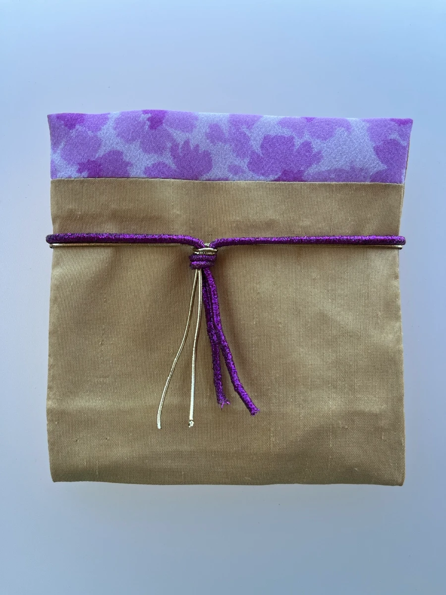 Sobre de tela dorado atado en el centro con un cordón morado brillante y dorado sobre un solapa con estampado floral lila.
