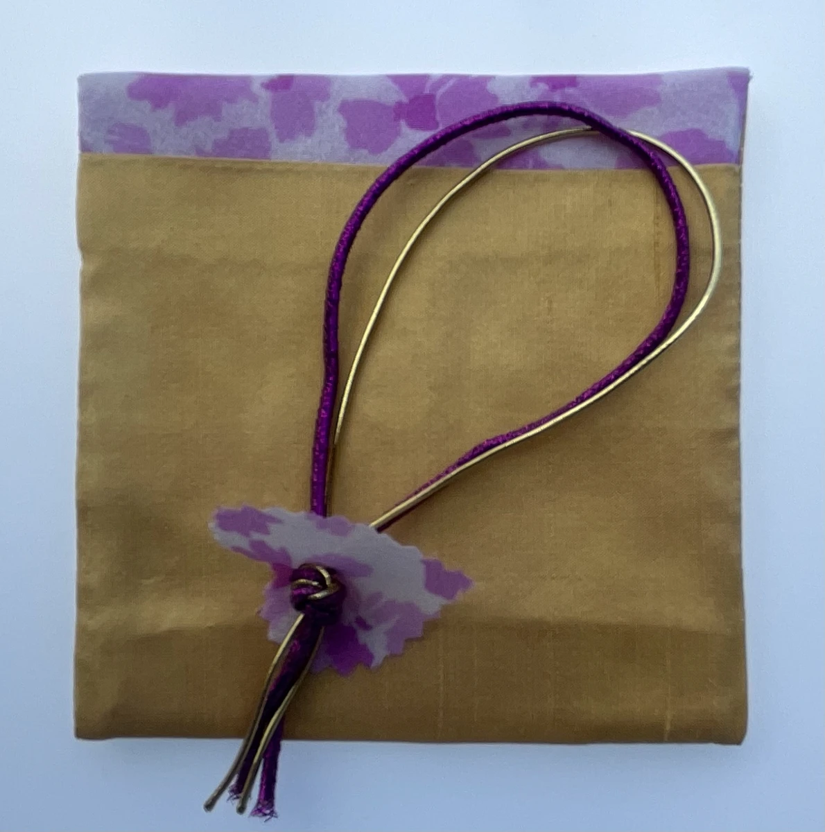 Paquete de tela dorado con cordón morado suelto y una flor decorativa de tela lila en el extremo del cordón.