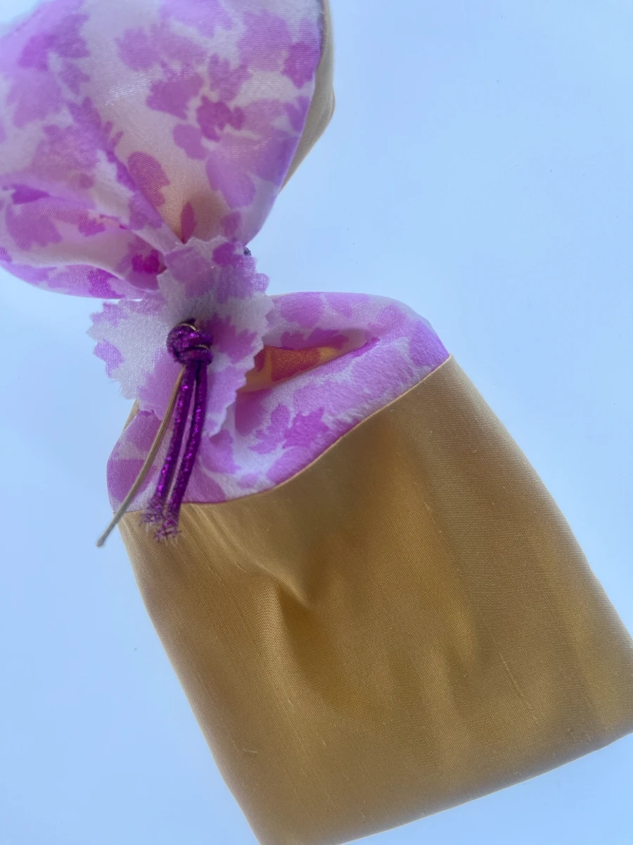 Saquito de regalo de tela dorada ajustado en la parte superior con un cordón morado, mostrando el forro interior de flores lilas.