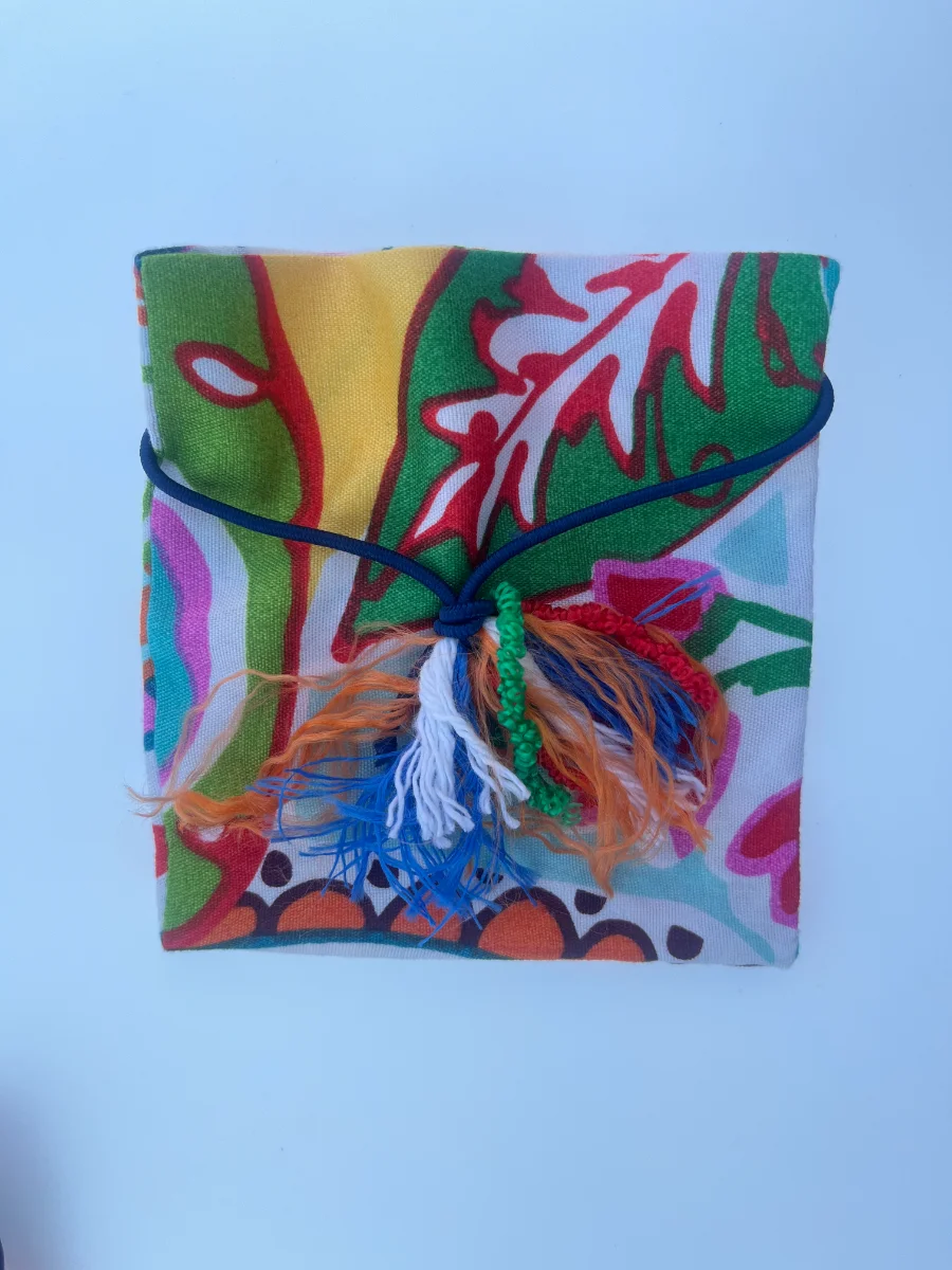 Envoltorio de tela tipo sobre con estampado vegetal en verde, amarillo y rojo, atado con una goma azul y una borla artesanal de hilos multicolor.