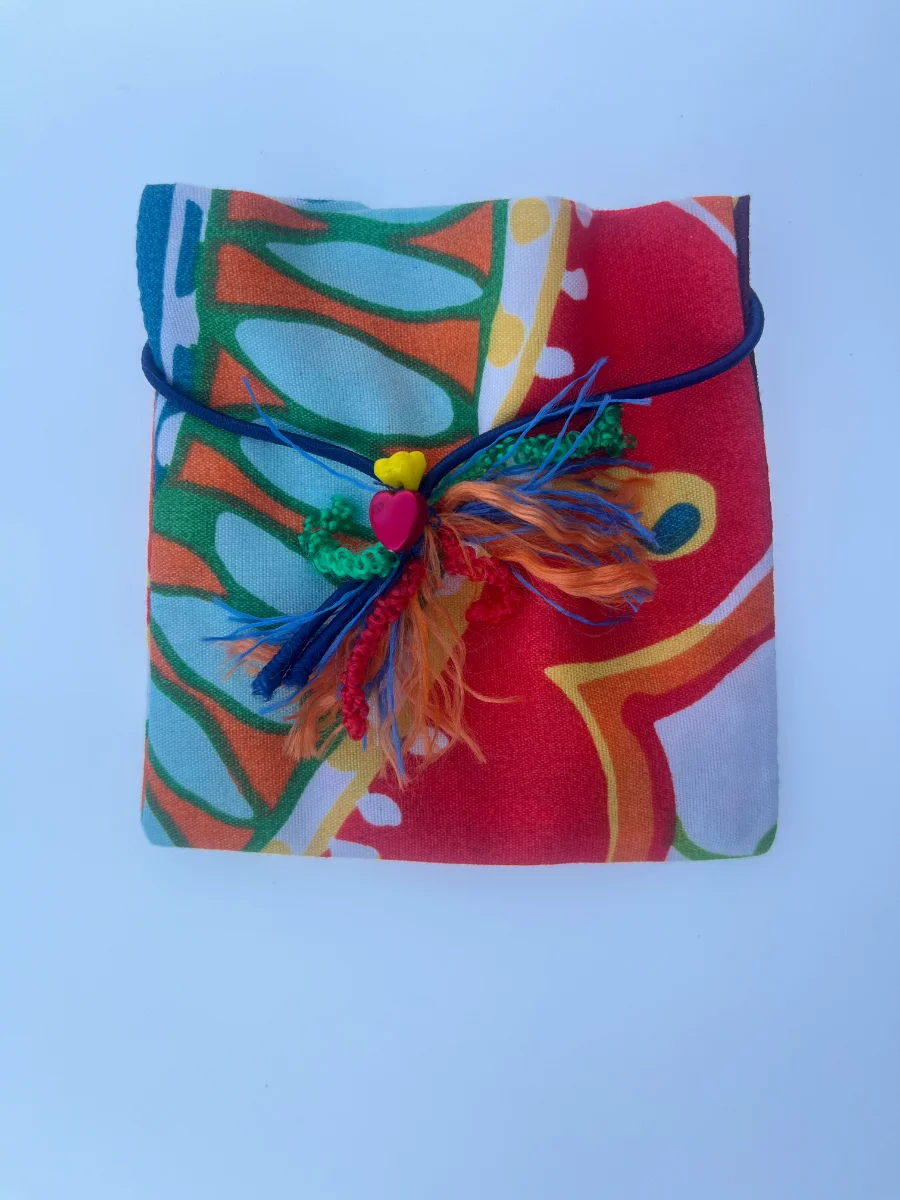 Paquete de tela tipo sobre en tonos rojos y naranjas, atado con cordón azul y rematado con una flor de hilos y un adorno de corazón rosa.