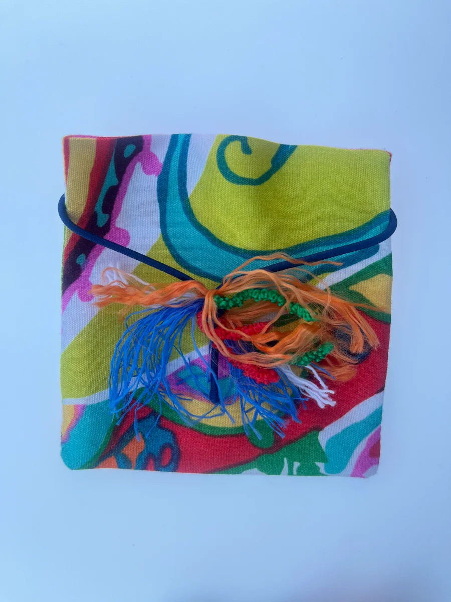 Sobre cuadrado de tela con diseño abstracto en amarillo, verde y azul, atado horizontalmente con goma azul y borla de hilos de colores.