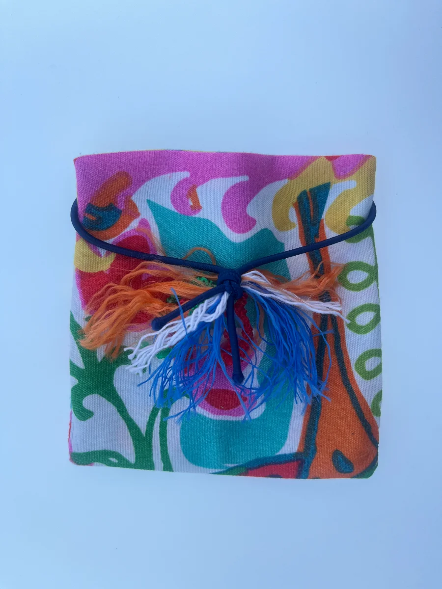 Paquete plano de tela con estampado de formas curvas en rosa, turquesa y naranja, ajustado con goma azul y flecos multicolores.
