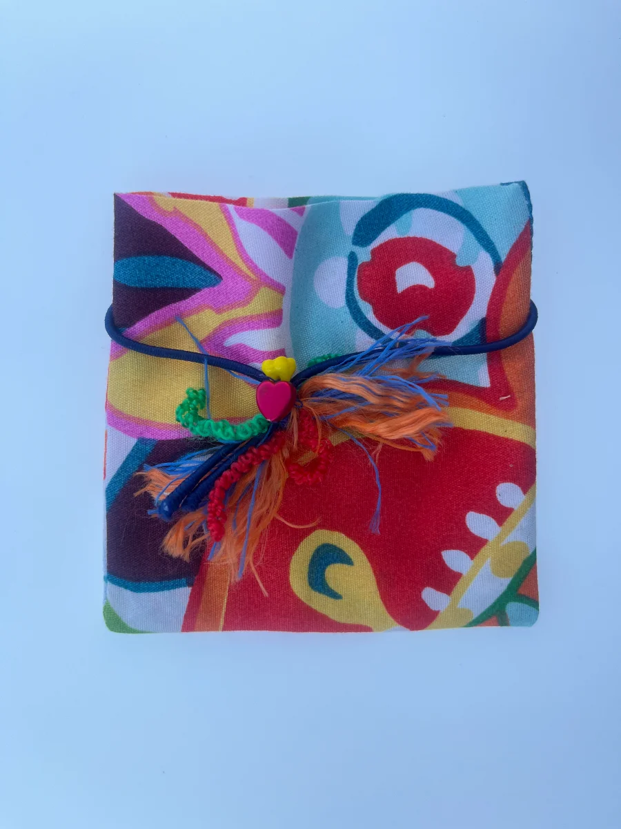 Sobre de tela con estampado rojo y amarillo, cerrado con una goma azul y un adorno central de corazón magenta con hilos.