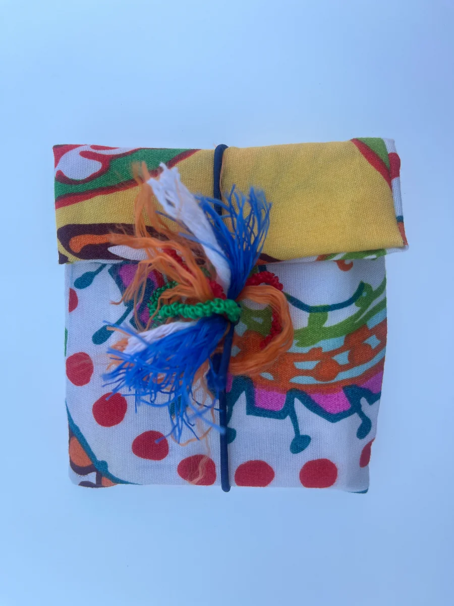Regalo envuelto verticalmente en tela de lunares rojos y solapa amarilla superior, sujeto por una goma vertical con borla multicolor.