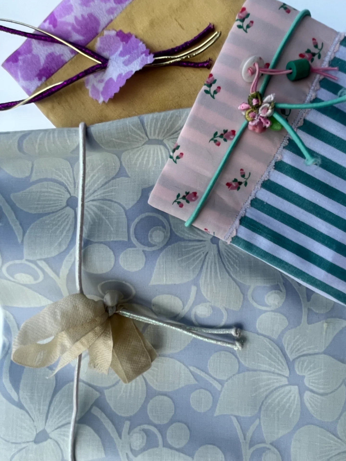 Vista superior de tres paquetes envueltos con distintas telas: azul floral, dorado con morado y franjas verdes con flores rosas.