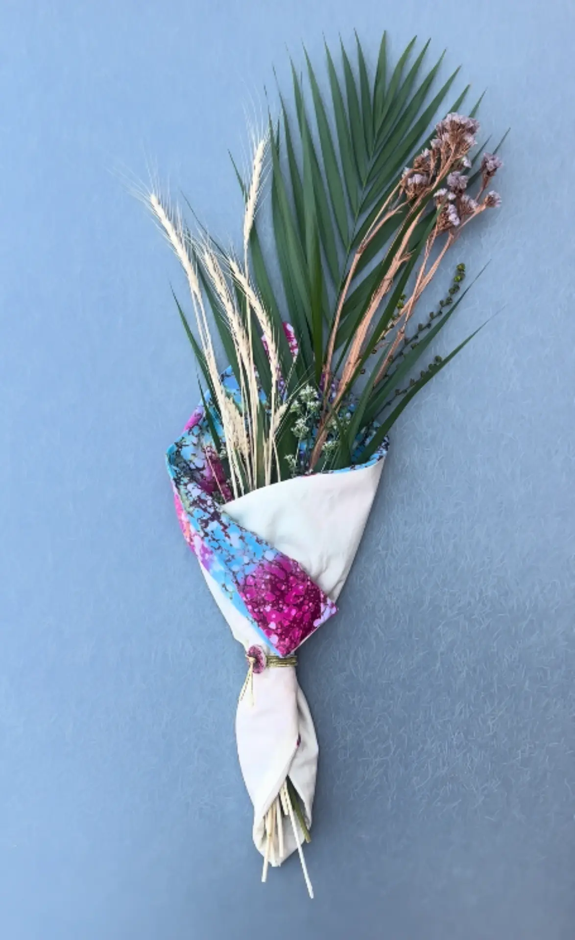 Ramo de espigas, hojas verdes y flores secas envuelto cuidadosamente en una tela artesanal multicolor y atado con un cordón.