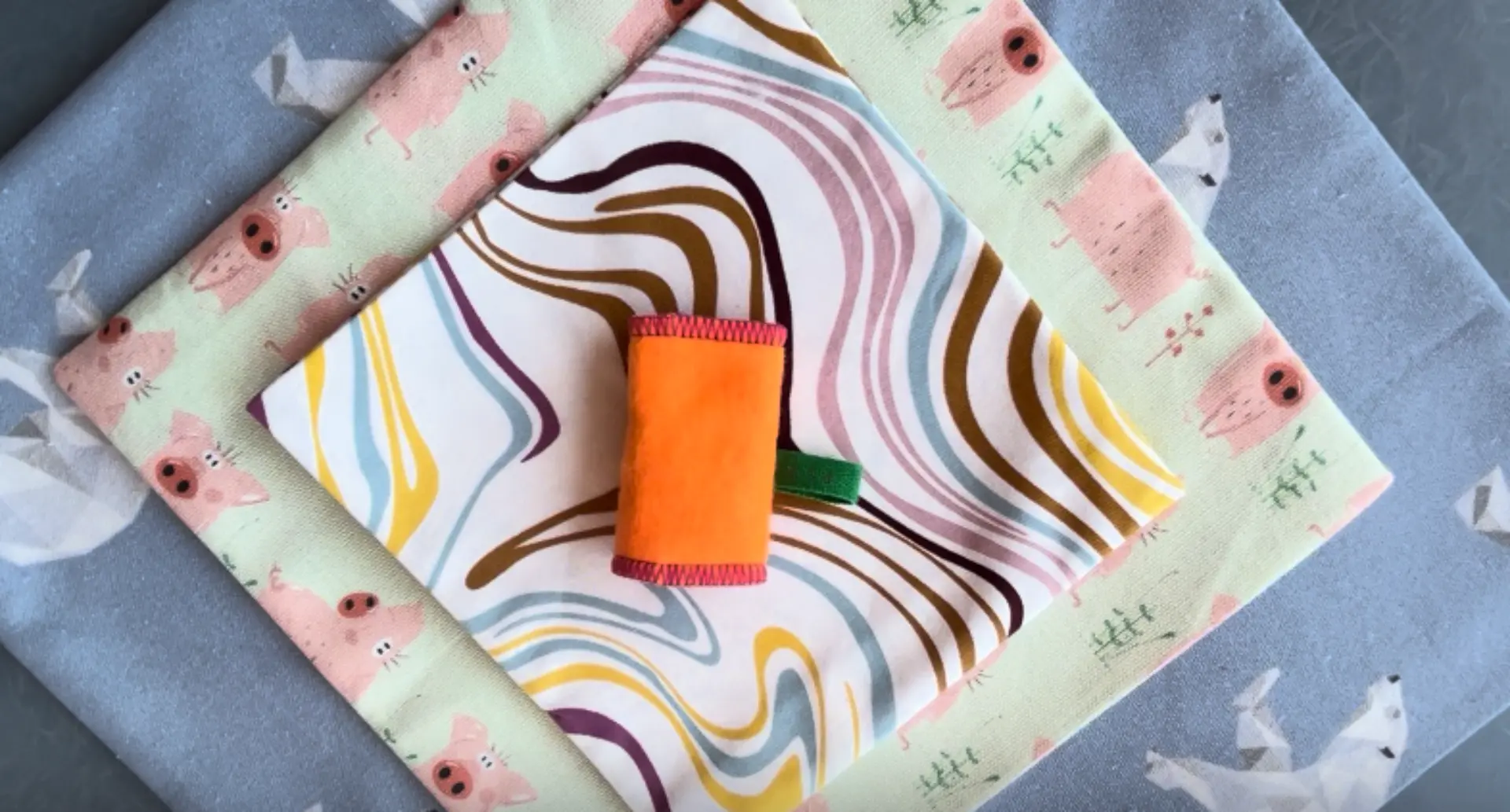 Tres telas estampadas superpuestas con un pequeño rollo o estuche de tela naranja ubicado justo en el centro.